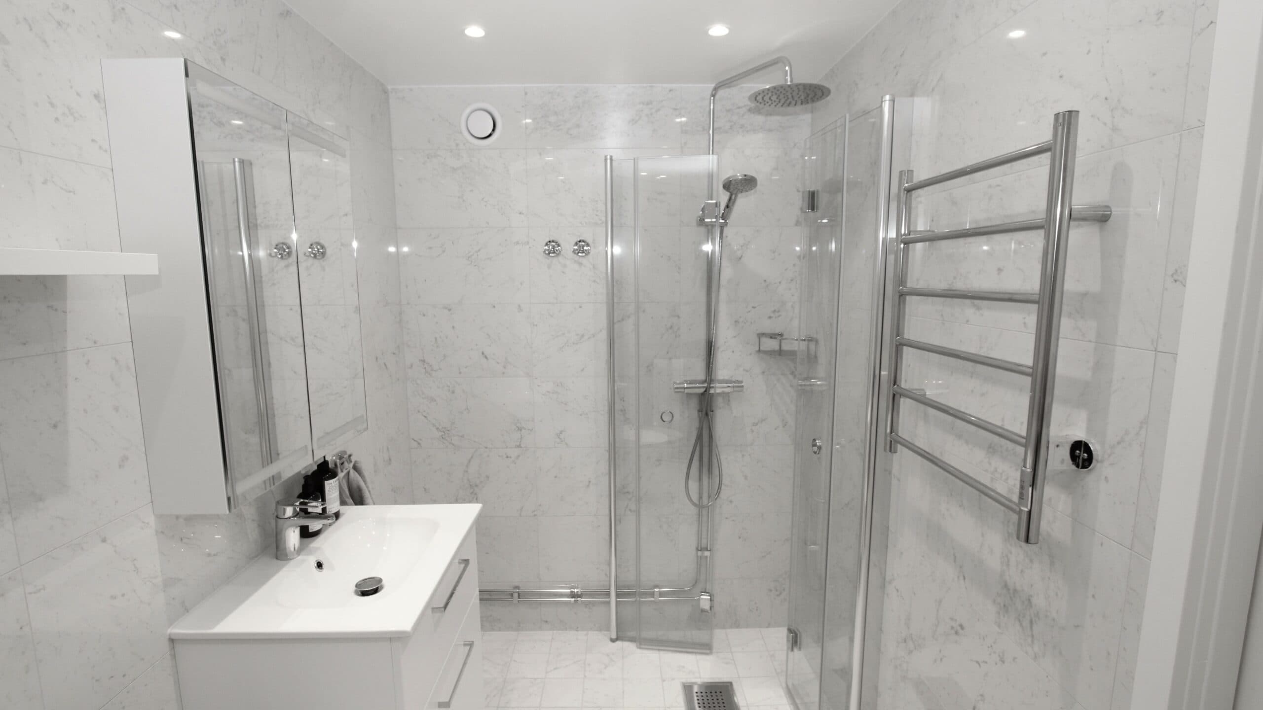 Modernt badrum med ljusa kakelväggar, vägghängd toalett och dusch med glasvägg samt svart armatur.