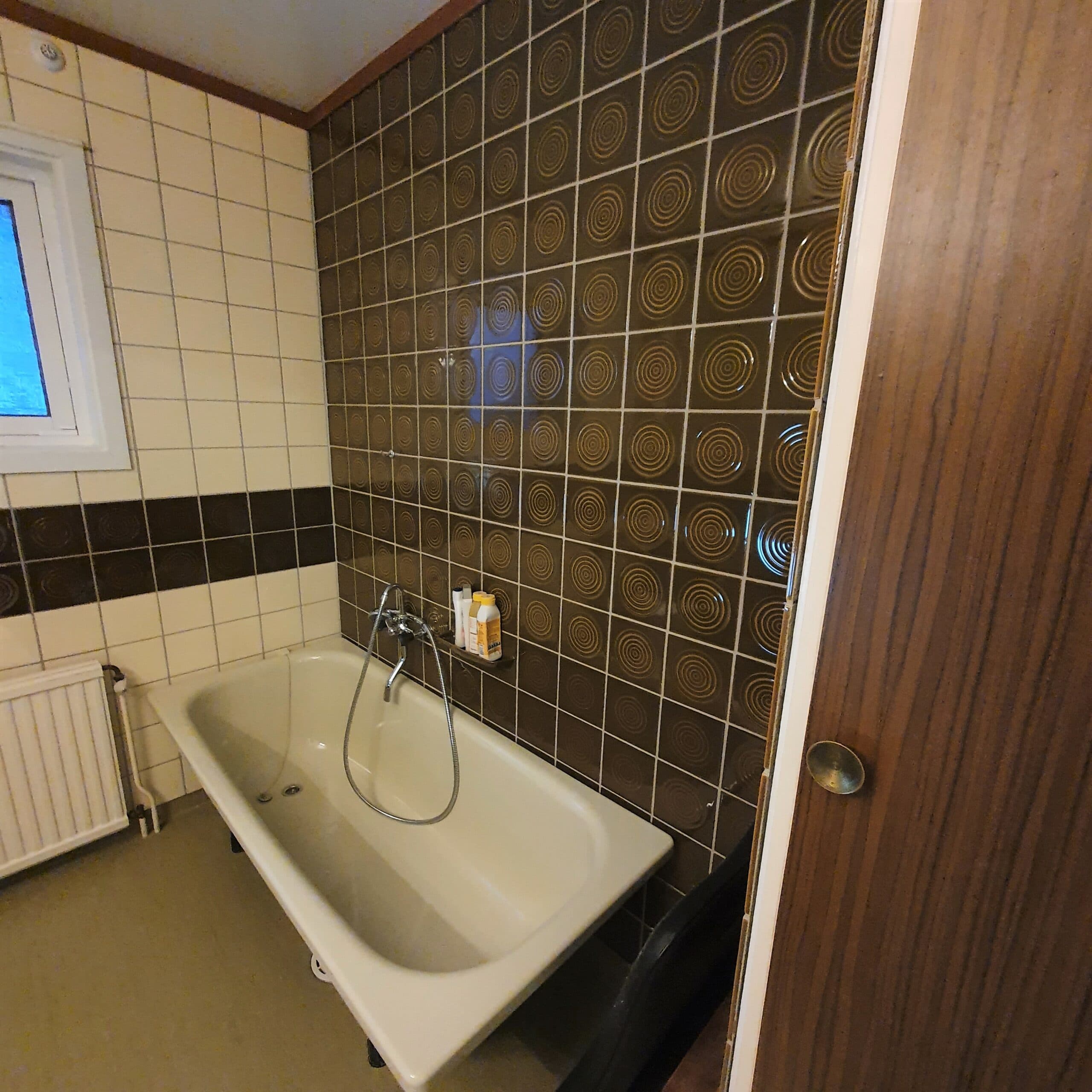 Ljust badrum med fristående badkar, vita kakelväggar, fönster med naturligt ljusinsläpp och trädörr